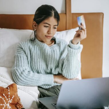 A young woman relaxes in bed, shopping online with a laptop and credit card in hand.